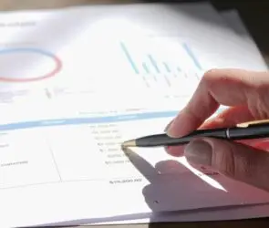Hand holding a pen reviewing a printed design business budget sheet with graphs and charts.
