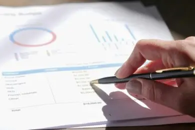 Hand holding a pen reviewing a printed design business budget sheet with graphs and charts.