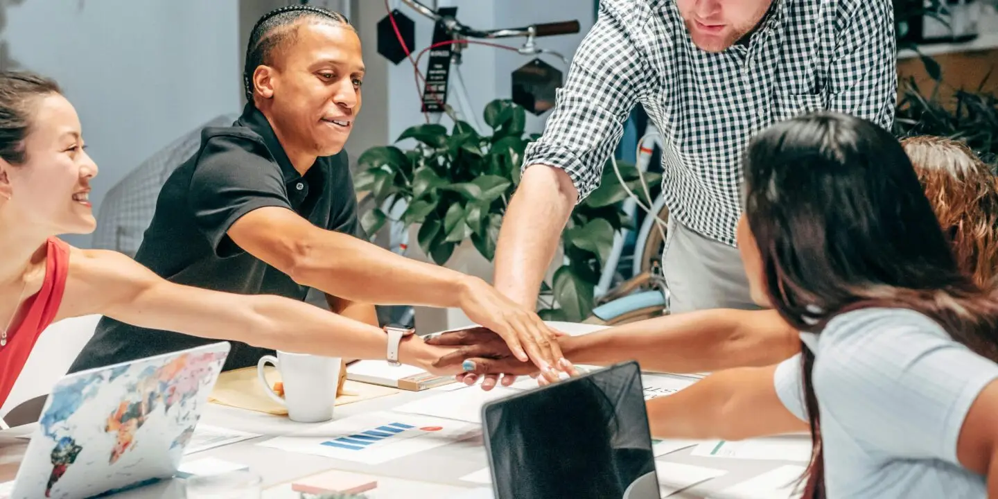 Diverse creative design team joining hands in a collaborative meeting around a shared workspace.