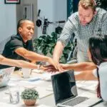 Diverse creative design team joining hands in a collaborative meeting around a shared workspace.