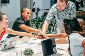 Diverse creative design team joining hands in a collaborative meeting around a shared workspace.