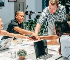 Diverse creative design team joining hands in a collaborative meeting around a shared workspace.