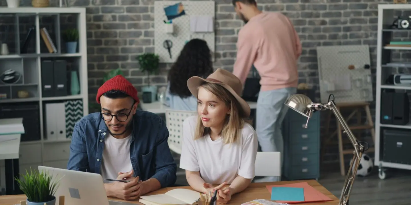 Designers collaborating in a creative design studio while reviewing marketing visuals and brand materials with AI on a laptop.