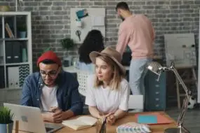 Designers collaborating in a creative design studio while reviewing marketing visuals and brand materials with AI on a laptop.