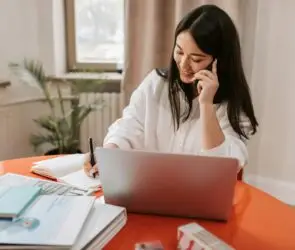 Smiling design business woman taking notes during a phone call with a client while working on her laptop.