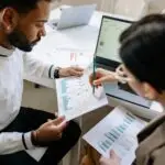 Design entrepreneur analyzing printed charts and graphs with a laptop to assess financial growth and business performance.