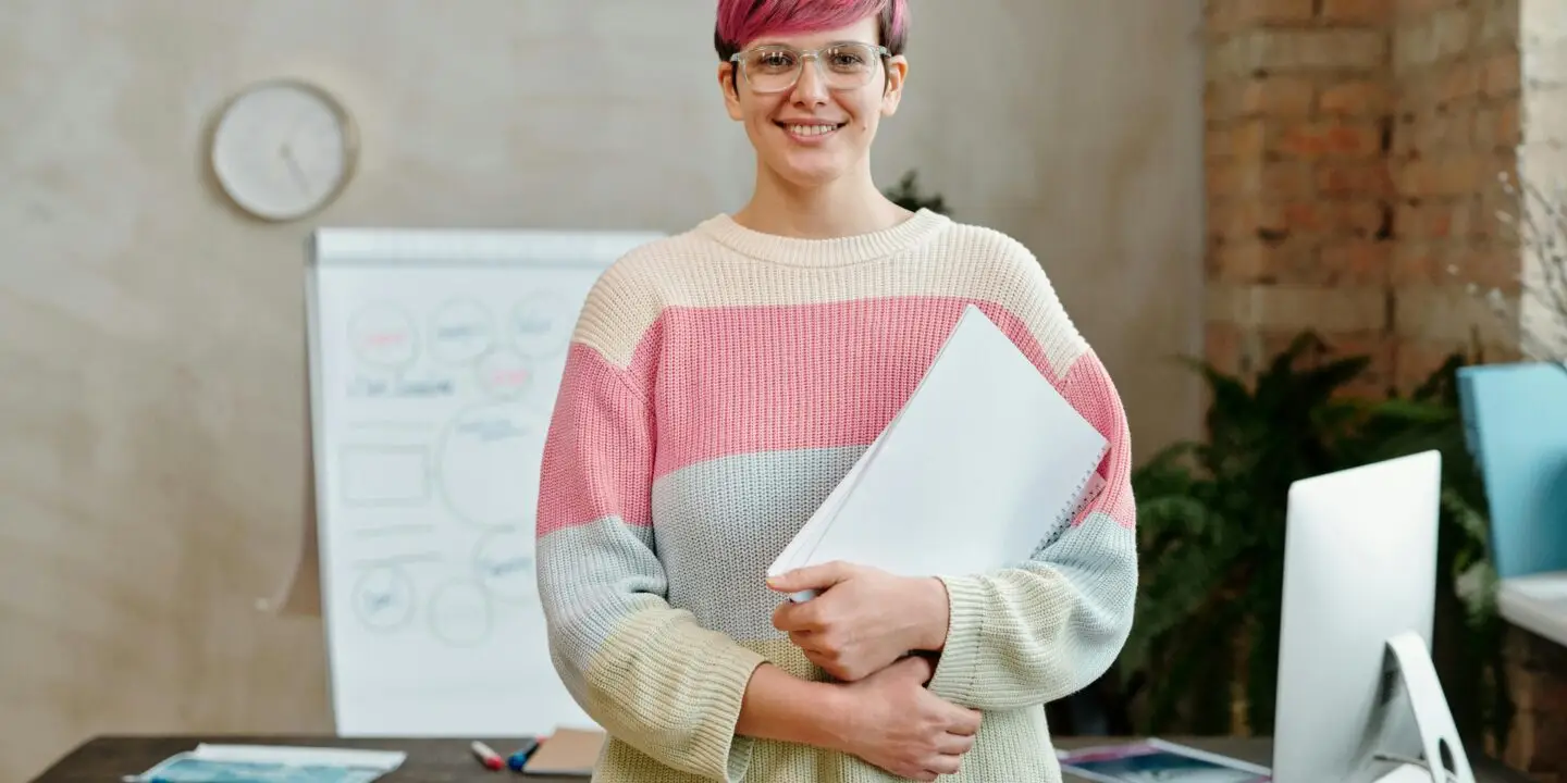 Confident freelance designer standing in a modern creative workspace, holding a notebook with design strategy plans, symbolizing business growth and scaling success.