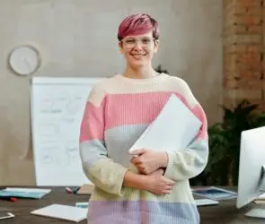 Confident freelance designer standing in a modern creative workspace, holding a notebook with design strategy plans, symbolizing business growth and scaling success.