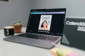 Laptop displaying a Canva-designed 2025 personal planner template on a desk with coffee, plant, and calendar board.