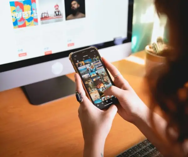 Design entrepreneur managing digital content and social media on smartphone while working at desktop computer.