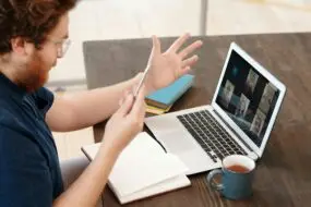 Designer participating in a video conference on a laptop while holding a smartphone at a desk.