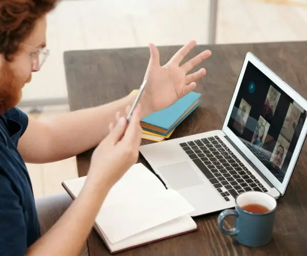 Designer participating in a video conference on a laptop while holding a smartphone at a desk.
