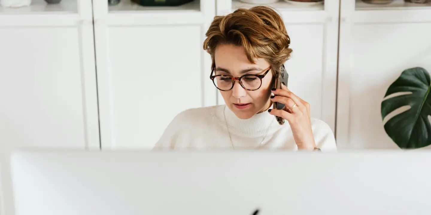 Woman talking on phone while working behind a desktop computer in a home office.