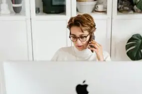 Woman talking on phone while working behind a desktop computer in a home office.
