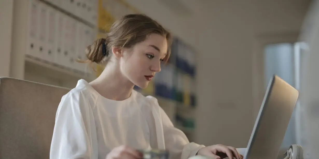 Woman working on laptop at desk while holding a payment card in a home office.