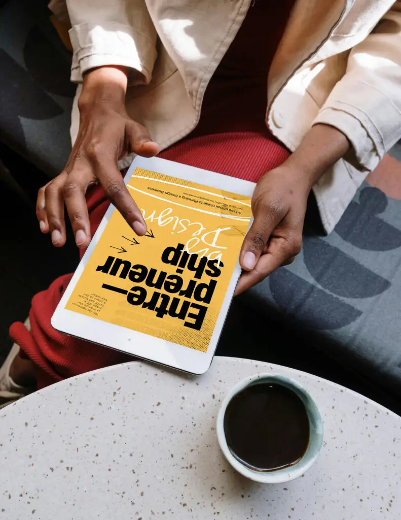 Person holding a tablet displaying the “Entrepreneurship by Design” digital guide while sitting near a table with coffee.