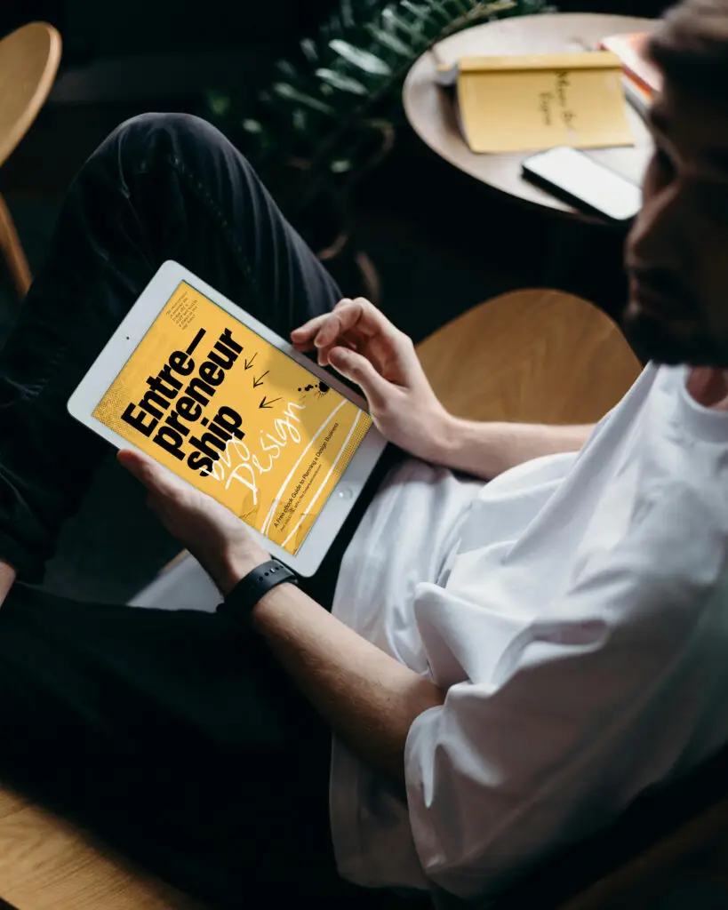 Man sitting in a chair holding a tablet displaying the “Entrepreneurship by Design” digital guide on screen.