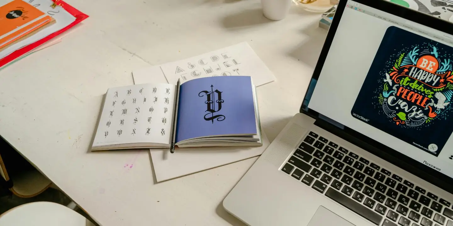 Designer workspace featuring typography references, lettering designs, and a laptop displaying digital artwork creation.