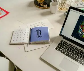 Designer workspace featuring typography references, lettering designs, and a laptop displaying digital artwork creation.