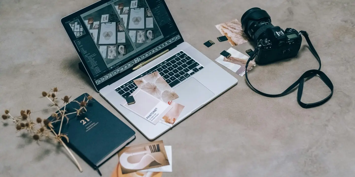 Laptop with creative design layouts next to a camera, memory cards, and branding materials on a workspace floor.