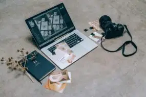 Laptop with creative design layouts next to a camera, memory cards, and branding materials on a workspace floor.