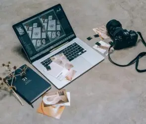 Laptop with creative design layouts next to a camera, memory cards, and branding materials on a workspace floor.