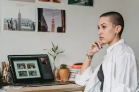 Graphic designer reflecting at her workspace while reviewing creative work on a laptop after a professional setback.