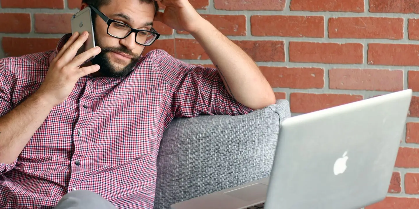 Freelance designer on a client phone call while working on a laptop and managing unexpected project requests.