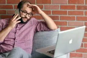 Freelance designer on a client phone call while working on a laptop and managing unexpected project requests.