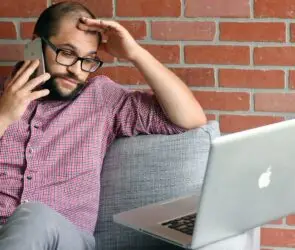 Freelance designer on a client phone call while working on a laptop and managing unexpected project requests.