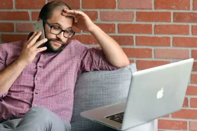 Freelance designer on a client phone call while working on a laptop and managing unexpected project requests.