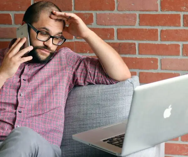 Freelance designer on a client phone call while working on a laptop and managing unexpected project requests.